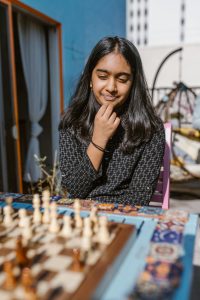 A young girl sits thoughtfully while playing chess outdoors in a bright setting.